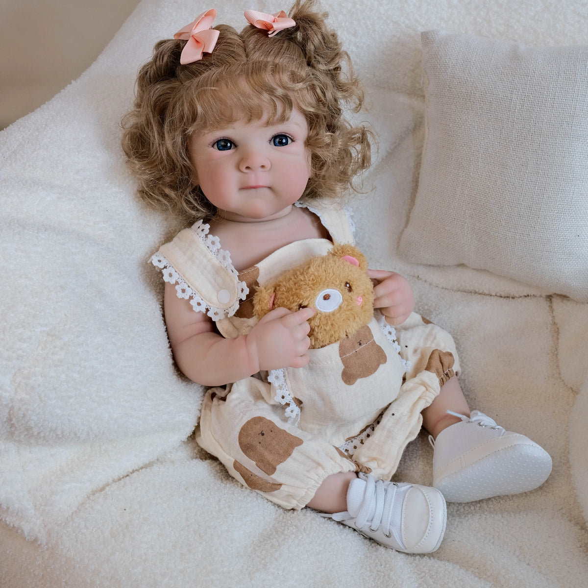 A lifelike 18-inch baby doll with hand-rooted hair wearing a light-colored outfit and sitting on a white blanket.