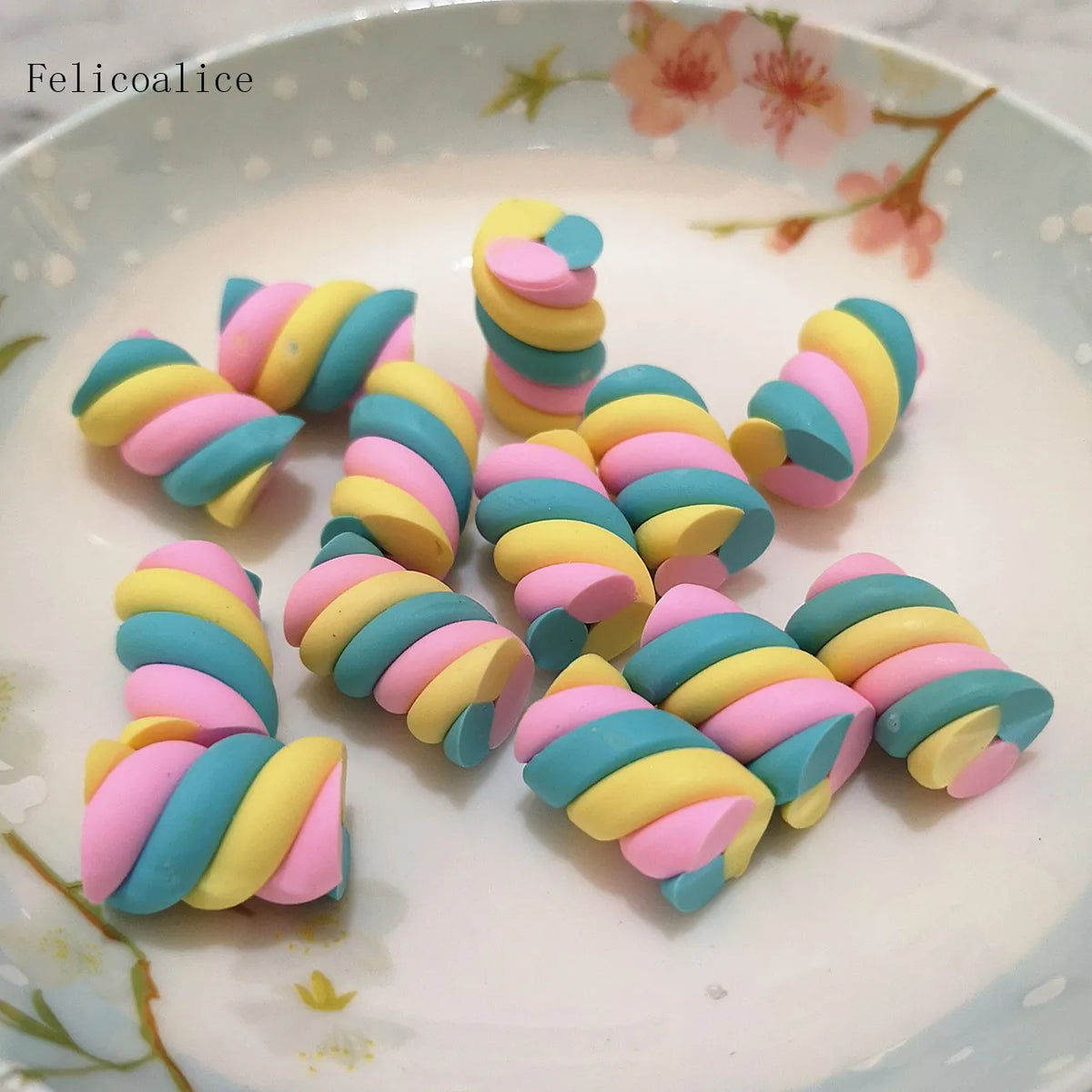 A collection of colorful polymer clay marshmallow charms in yellow, pink, and blue spiraled shapes arranged on a white plate.