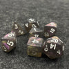 A set of seven iridescent acrylic polyhedral dice with a velvet pouch resting on a dark surface.