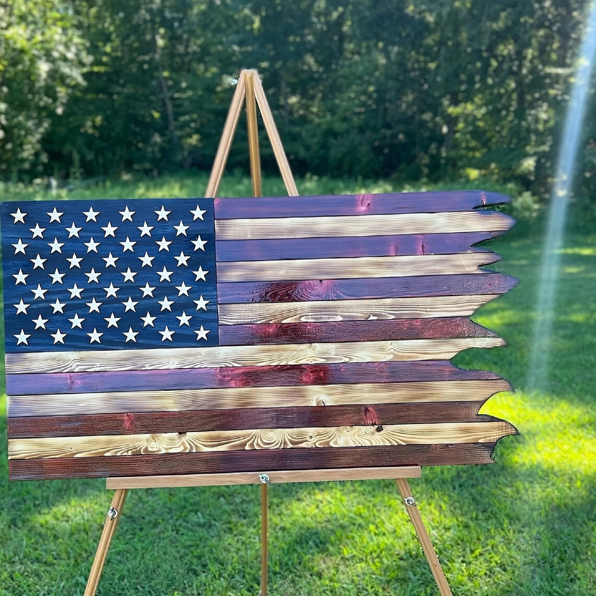 A wooden American flag with stars and stripes is displayed on a wooden stand in a grassy outdoor setting.