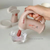A hand is holding a pink blender with a purple blade over a glass of water.