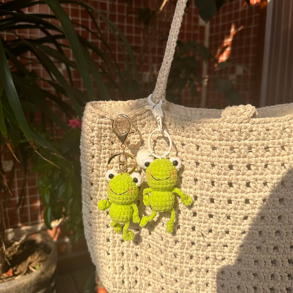 A handmade crochet frog keychain with bright green body and white eyes hanging from a cream-colored knitted bag.