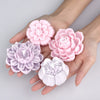 A hand holds several pink and purple flower-shaped candle wax pigment molds.