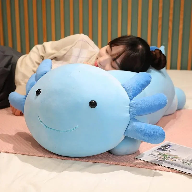 A light blue axolotl plush pillow is resting on a pink surface, with a person's face visible near it.