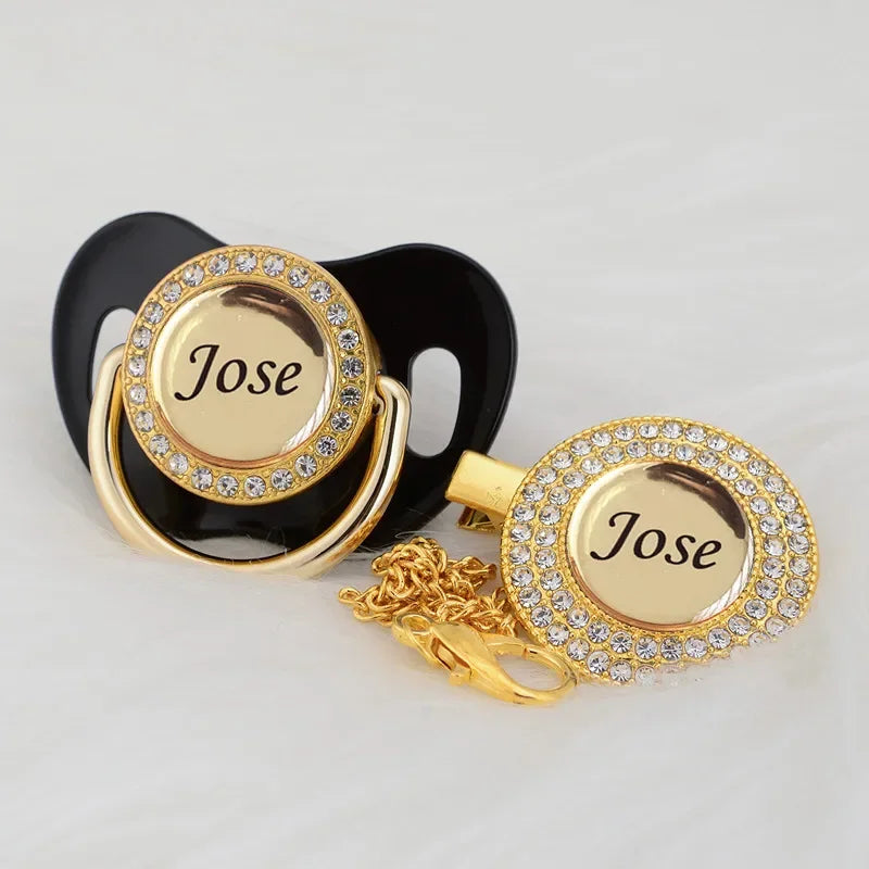 A gold and crystal pacifier clip set featuring a black silicone holder and a gold-toned ring with the name Jose engraved.