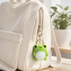 A beige fabric bag features a green crochet frog keychain attached to the zipper pull.