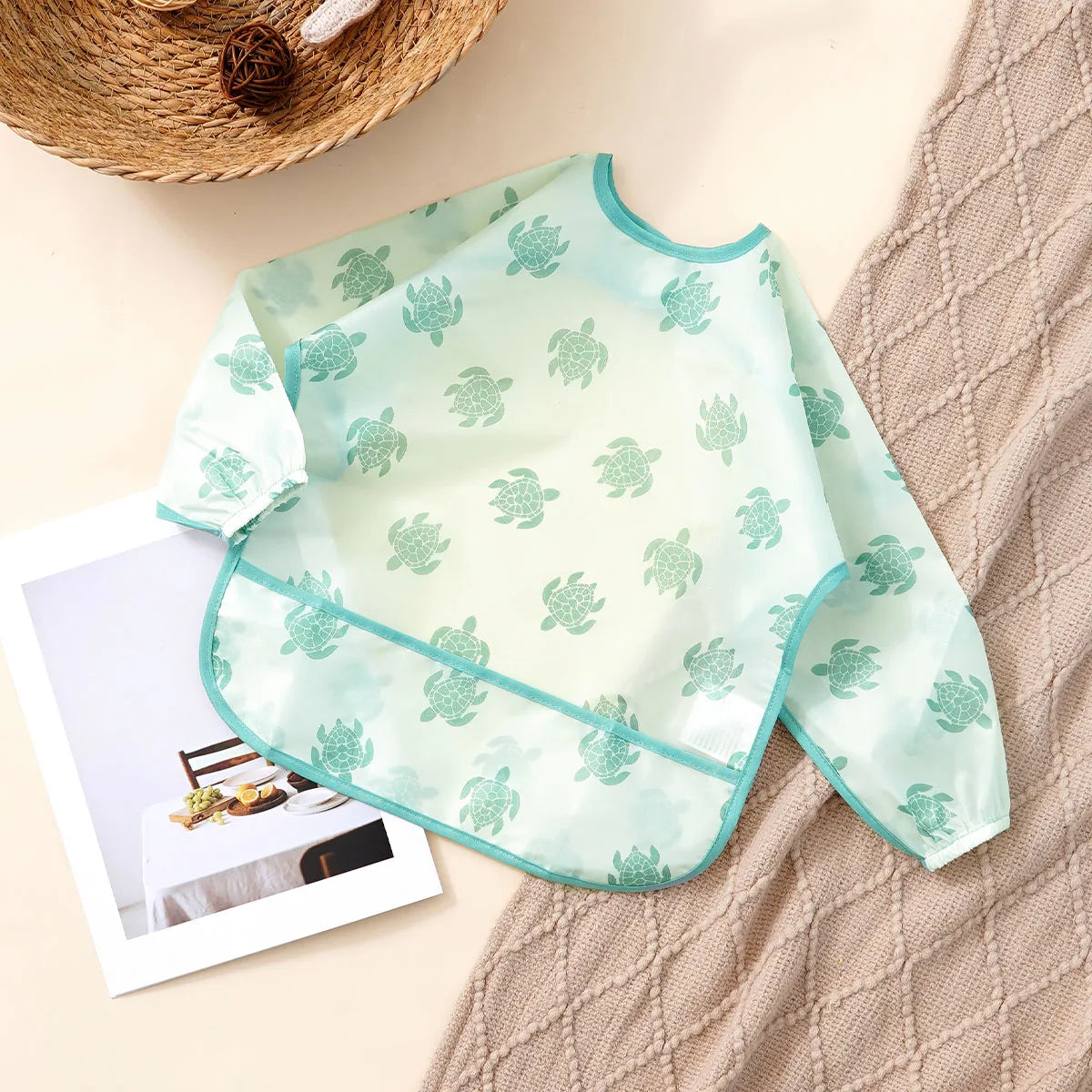 A light mint green waterproof long sleeve feeding bib with a subtle leaf pattern is displayed on a beige textured cloth.