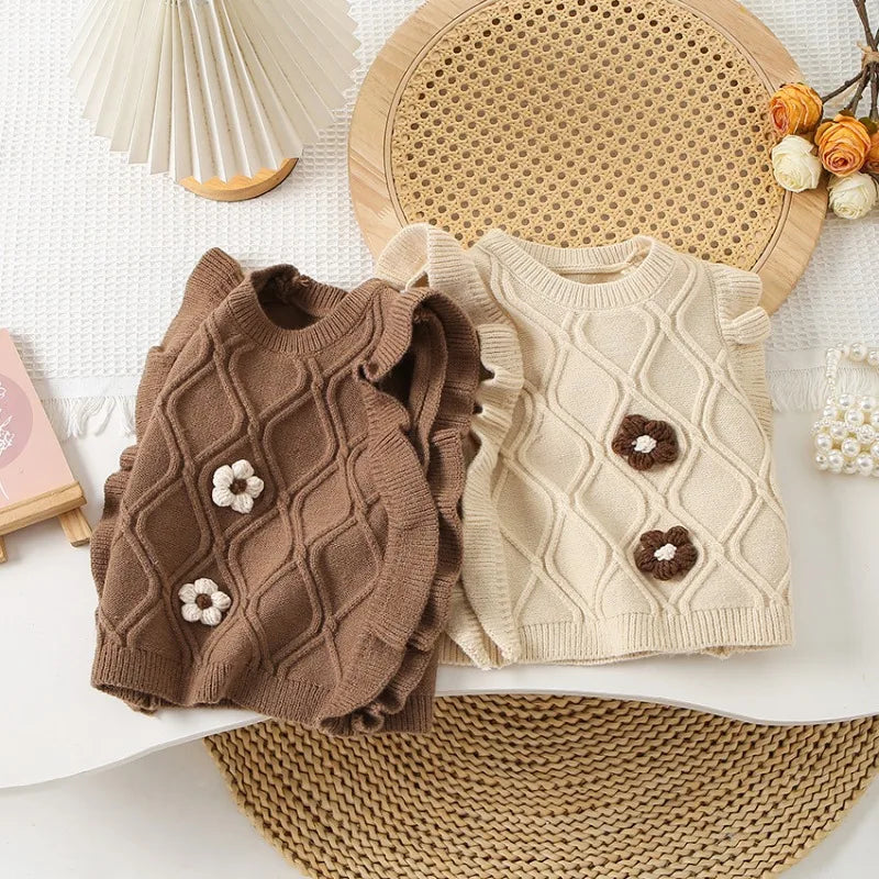 A brown knit sweater vest with a flower pattern and ruffled edge is displayed on a white surface next to a woven mat.