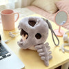 A soft gray dinosaur skeleton bag charm with a removable hood resting on a yellow table next to a laptop and a pink mug.