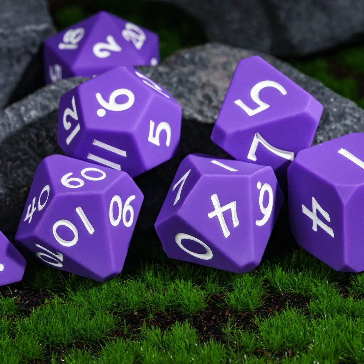 A collection of purple silicone polyhedral dice resting on green grass and dark stones.