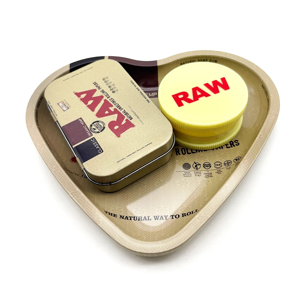 A love-shaped metal rolling tray holds a yellow herb grinder labeled RAW and a brown rolling paper.