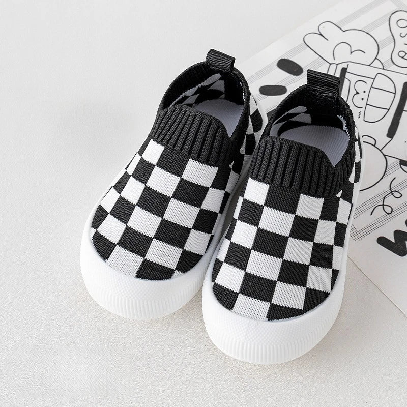 A pair of black and white checkered plaid slip-on toddler shoes for first walkers resting on a white surface.