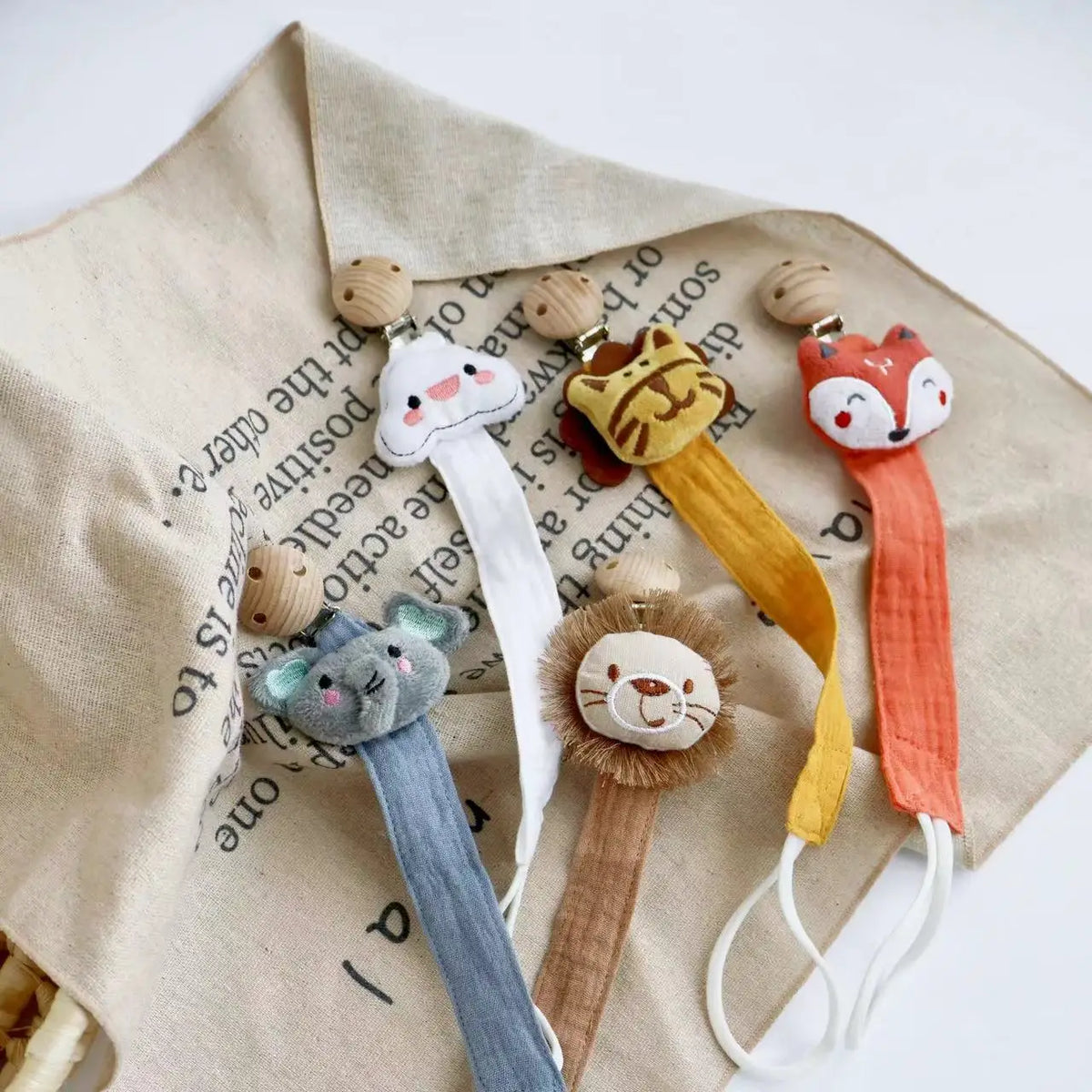 A set of elephant pacifier holders with clip in various colors like white, yellow, and orange on a beige fabric.