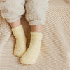 A pair of yellow knitted fleece socks worn by a person, resting on a beige knitted fabric.