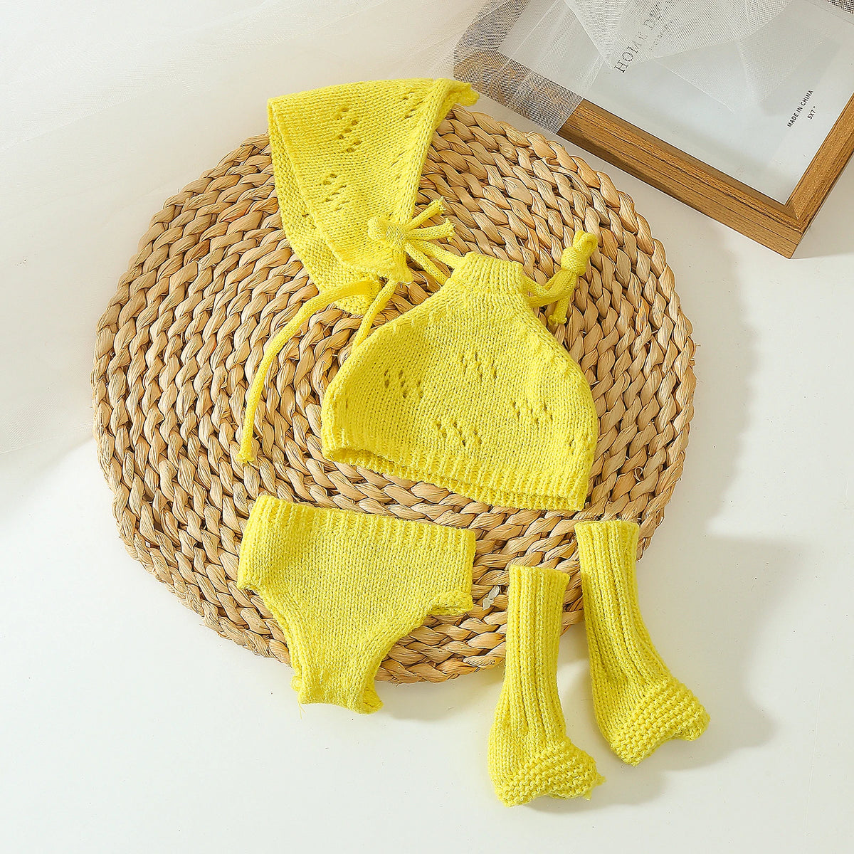 A yellow knitted doll outfit set featuring a hat, top, and three pairs of knitted socks on a woven mat.