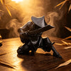 A dark 3D printed samurai warrior figurine with a hat and sword is displayed on a wooden surface amidst wisps of smoke.