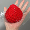 A bright red strawberry squeeze fidget toy is held in a person's hand, packaged in clear plastic.