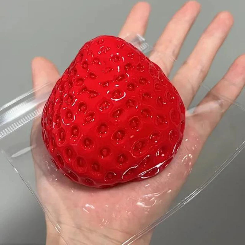 A bright red strawberry squeeze fidget toy is held in a person's hand, packaged in clear plastic.