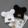 A pair of cozy knit baby beanie hats in white and black with black faux fur pom-poms.