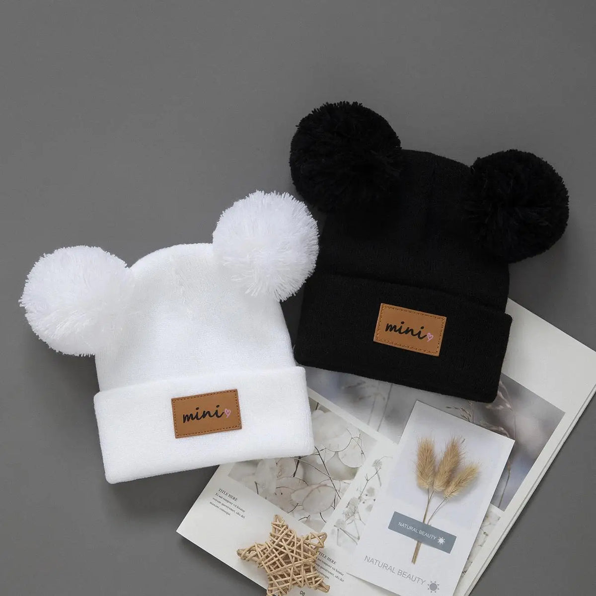 A pair of cozy knit baby beanie hats in white and black with black faux fur pom-poms.