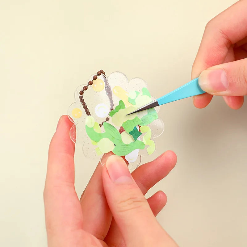 Hands use blue tweezers to apply gold and white glitter to a translucent sheet for nail art