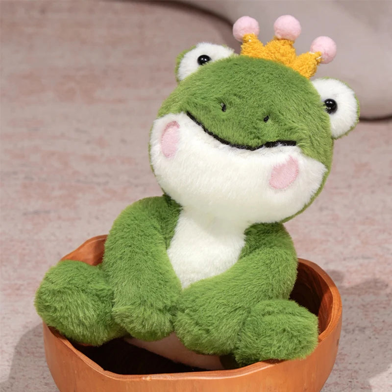 A cute green frog prince plush toy with a yellow crown sitting in a wooden bowl.
