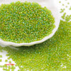 A pile of bright green glossy glass seed beads is displayed on a white dish and spilled onto a surface.
