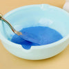 A spoon is scooping bright blue acrylic paint from a light blue bowl onto a neutral background.