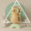 A soft, tan baby head protector pillow with ears and adjustable straps is displayed in front of a geometric green background.