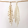 A set of two white baby gauze swaddle blankets with small brown animal patterns hanging from a wooden branch.