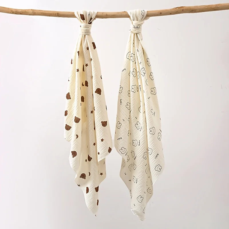 A set of two white baby gauze swaddle blankets with small brown animal patterns hanging from a wooden branch.