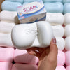 A hand holds a white soap ball squishy fidget toy with a pink box and other similar toys in the background.