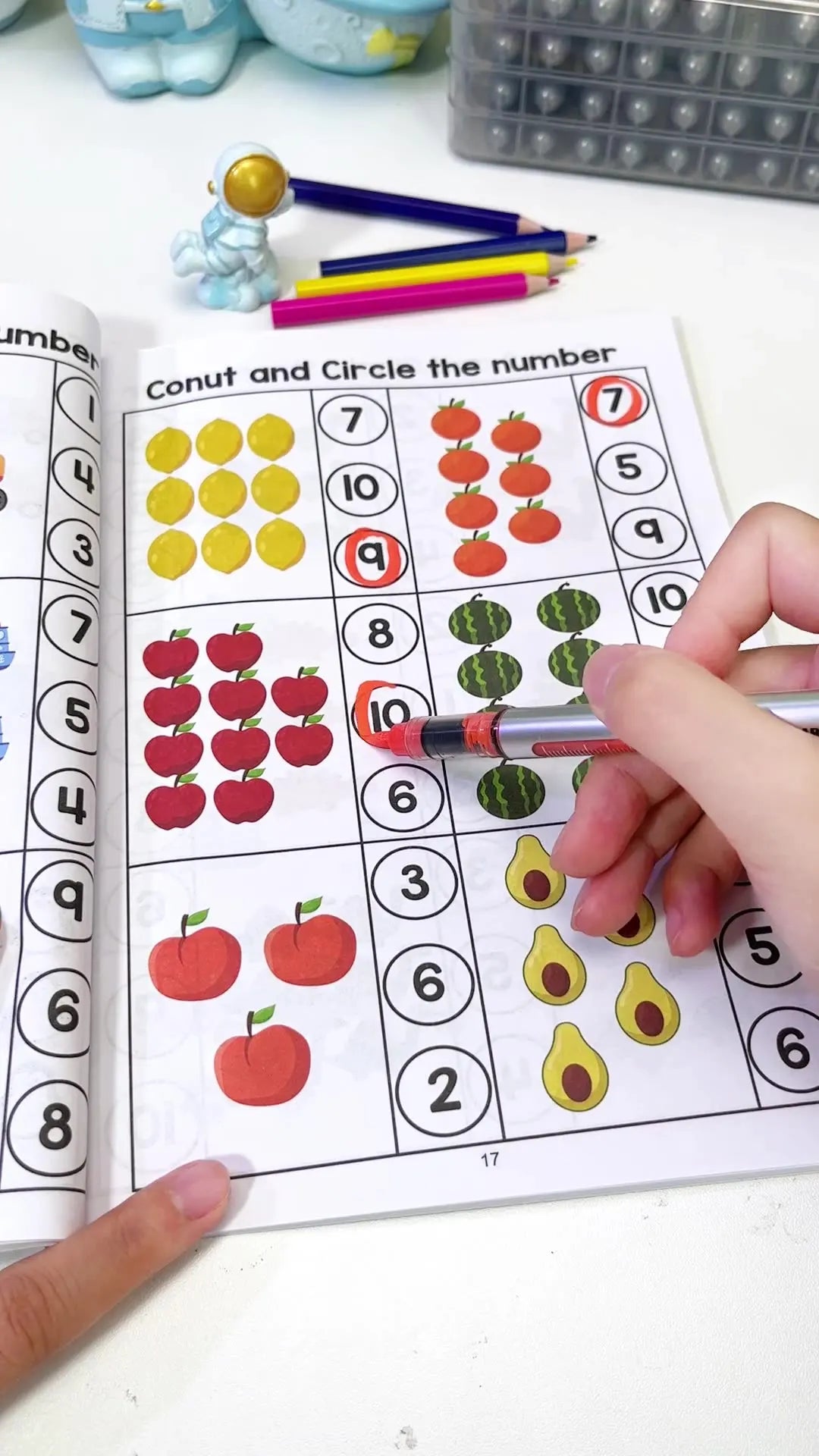 A child's hand is pointing to a preschool math workbook page with numbers and corresponding fruits for tracing and counting.