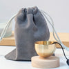 A gray fabric hand-shaped meditation pouch with white cord is shown next to a small brass singing bowl on a wooden base.