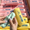 A hand holds a yellow squeeze butter stress relief toy next to a stack of pink and yellow butter blocks.