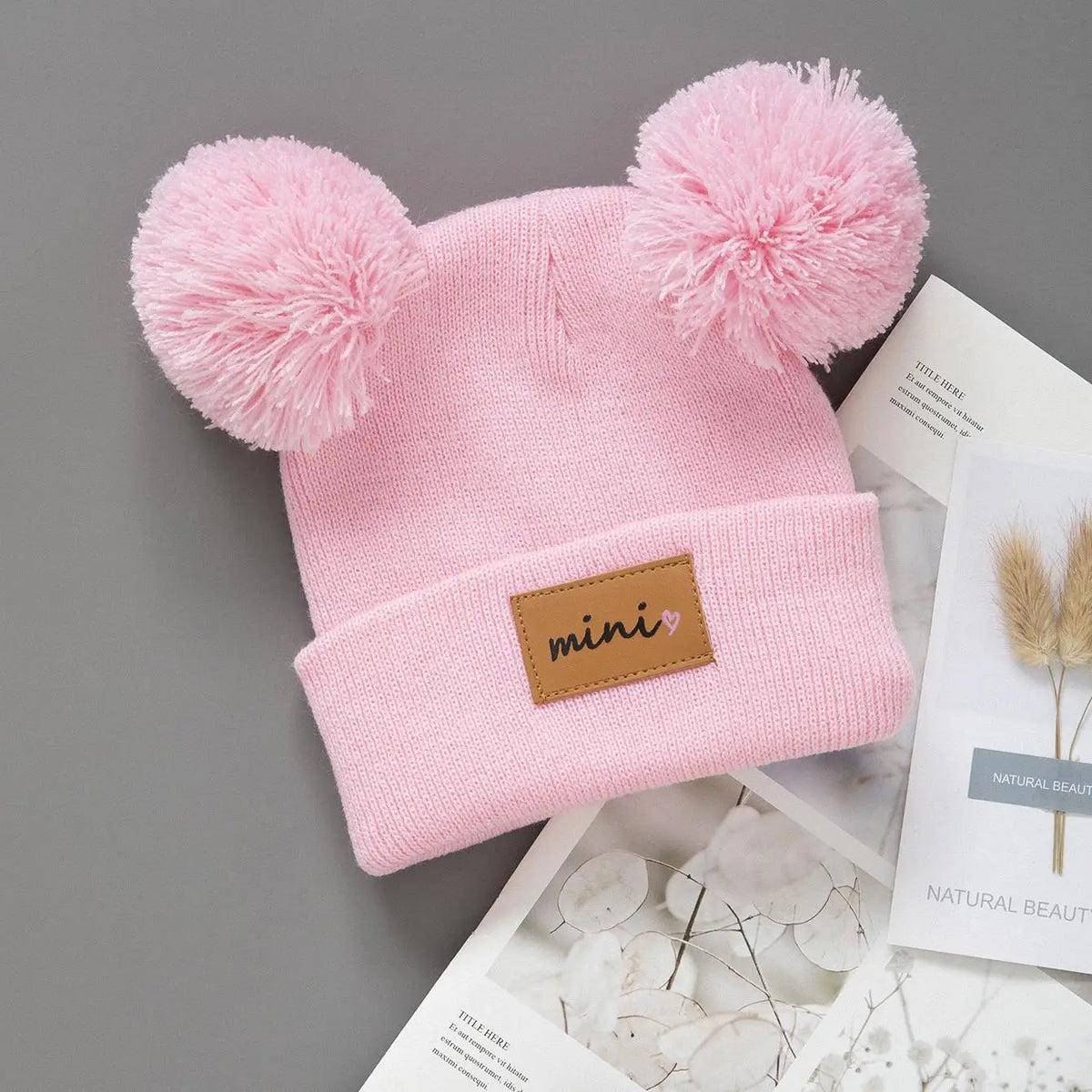 Pink knitted baby beanie hat with fluffy pink pom-poms and a brown label