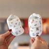 A pair of white soft sole sandals for a baby, featuring bear-themed fabric and small grey animal appliqués.