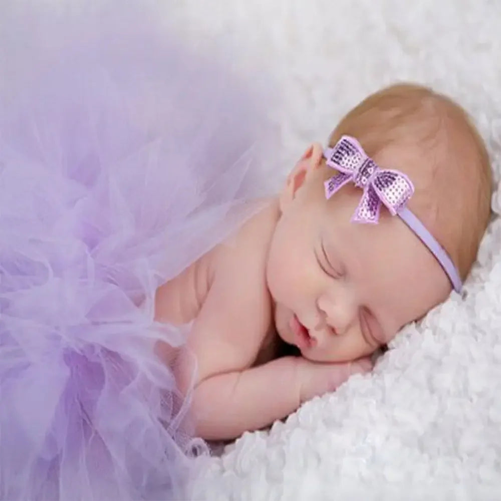 A sleeping baby girl wearing a purple tutu dress and a matching purple bow headband, resting on white fluffy material.