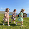 Three young girls in plaid dresses running across a grassy field near the sea