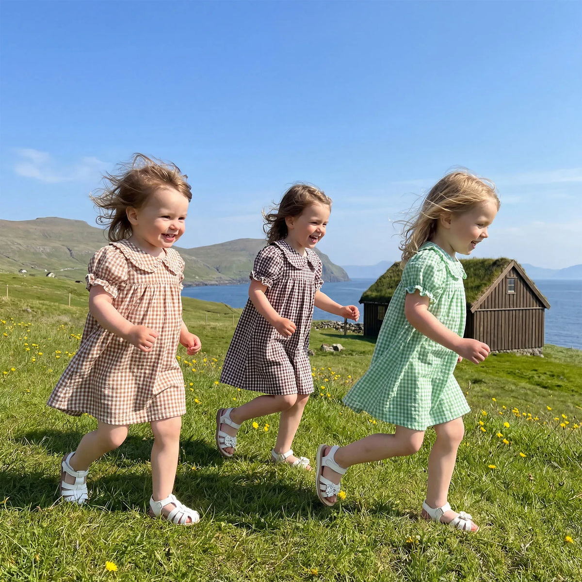 Three young girls in plaid dresses running across a grassy field near the sea