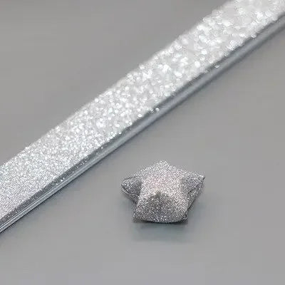 Silver glitter origami star paper strips laid diagonally on a gray surface.