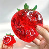 A hand holds a bright red squishy stress ball shaped like a strawberry with green leaves.