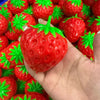A hand holds a bright red strawberry crunchy stress ball with green leaves, surrounded by other similar toys.
