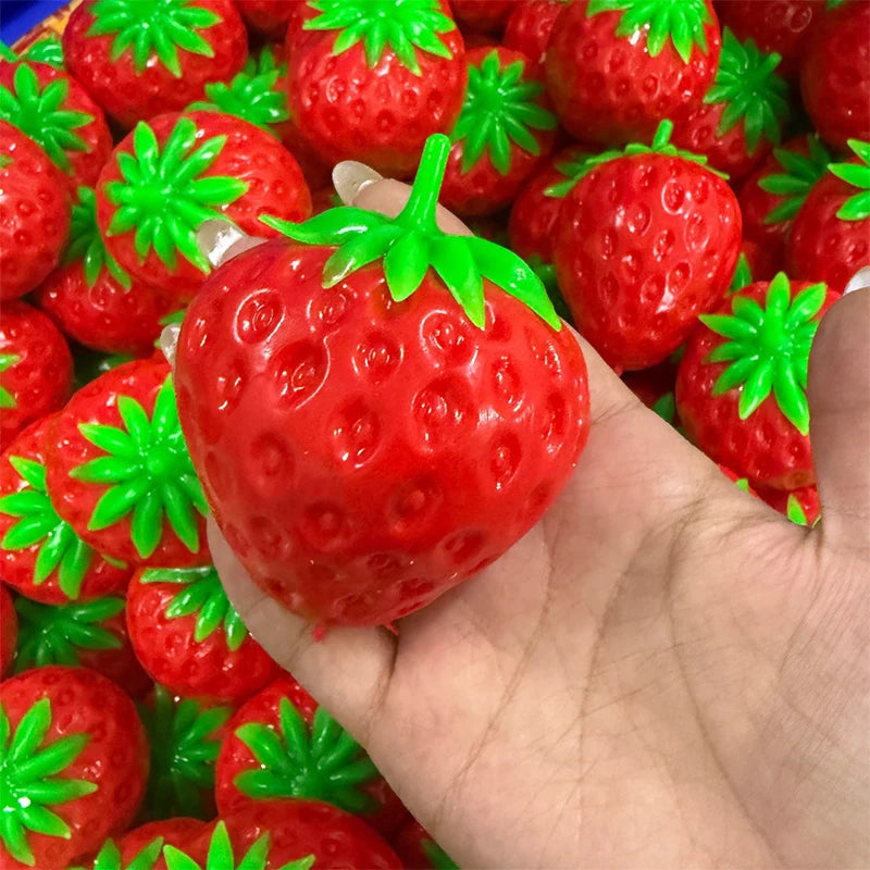A hand holds a bright red strawberry crunchy stress ball with green leaves, surrounded by other similar toys.