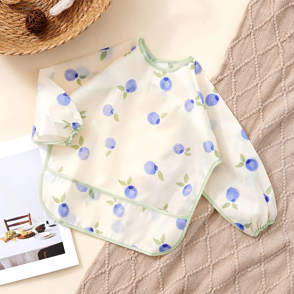 A white waterproof long sleeve feeding bib with a light blue blueberry pattern lying on a beige textured cloth.