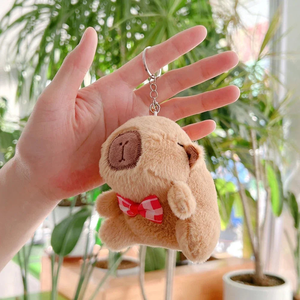A hand holds an adorable capybara plush keychain charm in a bright indoor setting.