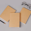 A stack of blank kraft paper notebooks and a black pen are displayed on a gray surface.