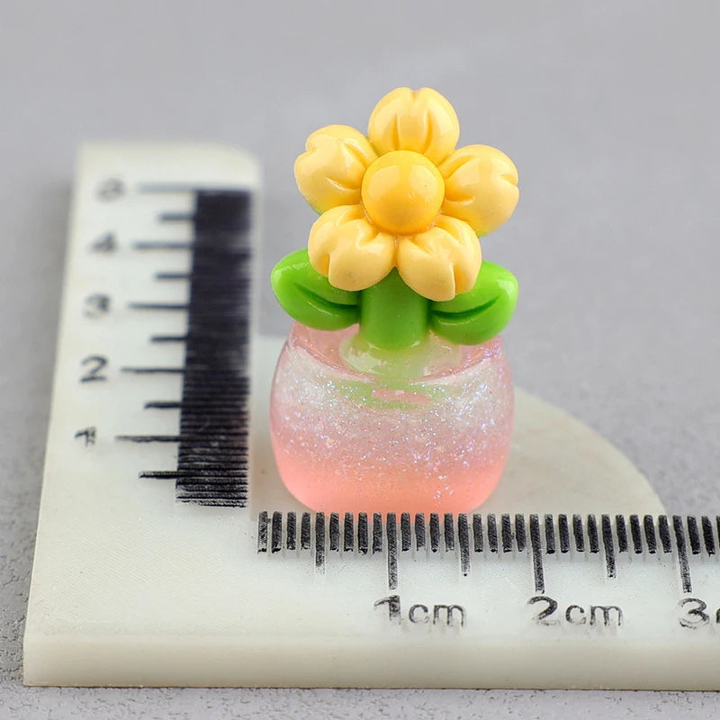 A miniature yellow flower on a green stem sits in a pink glittery glass pot, measured against a ruler.