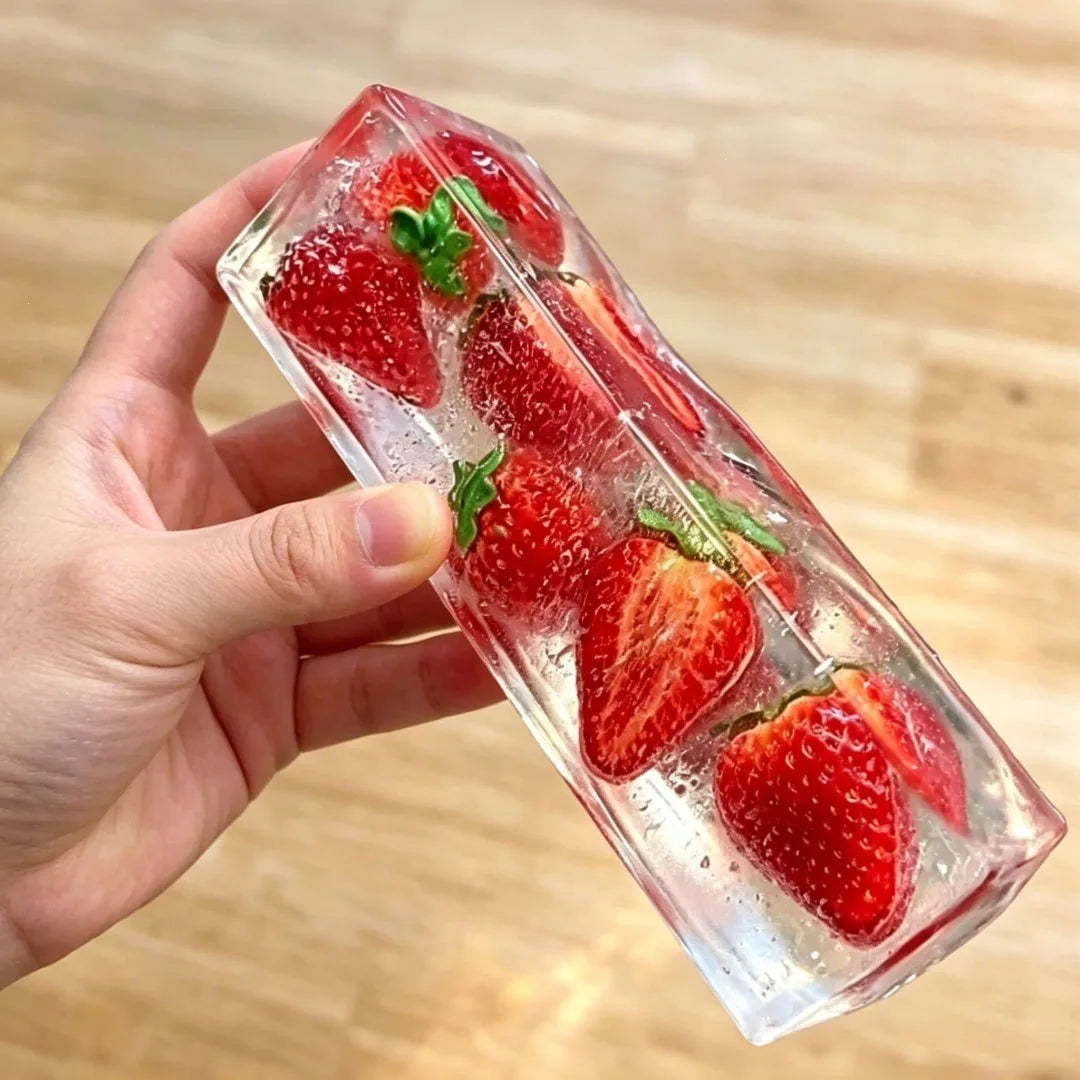 A hand holds a clear ice cube filled with fresh strawberries and mint leaves.