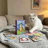 A white cat sits on a gray blanket surrounded by various tarot cards.